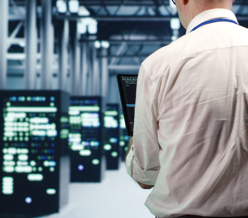 Aged IT consultant in data center, solving tasks on tablet while running scripts on laptop terminal. Trained repairman walking amidst servers, accessing databases and manipulating lines of code
